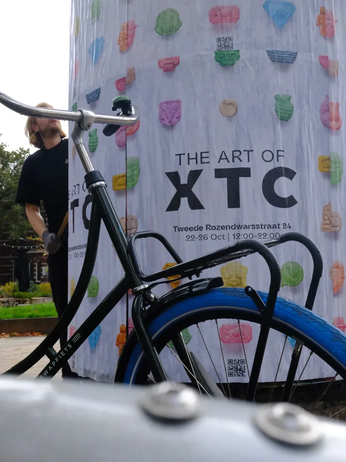 자전거 뒤 벽에 붙은 알약 그림 포스터. 「THE ART OF XTC」 「Tweede Rozendwarsstraat 24」 「22-26 Oct | 12:00-22:00」