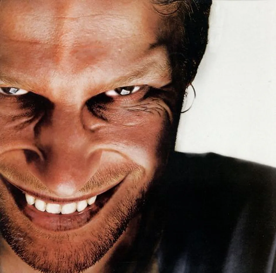 흰 배경 앞에서 이를 드러내고 기묘하게 웃는 Aphex Twin의 얼굴 클로즈업