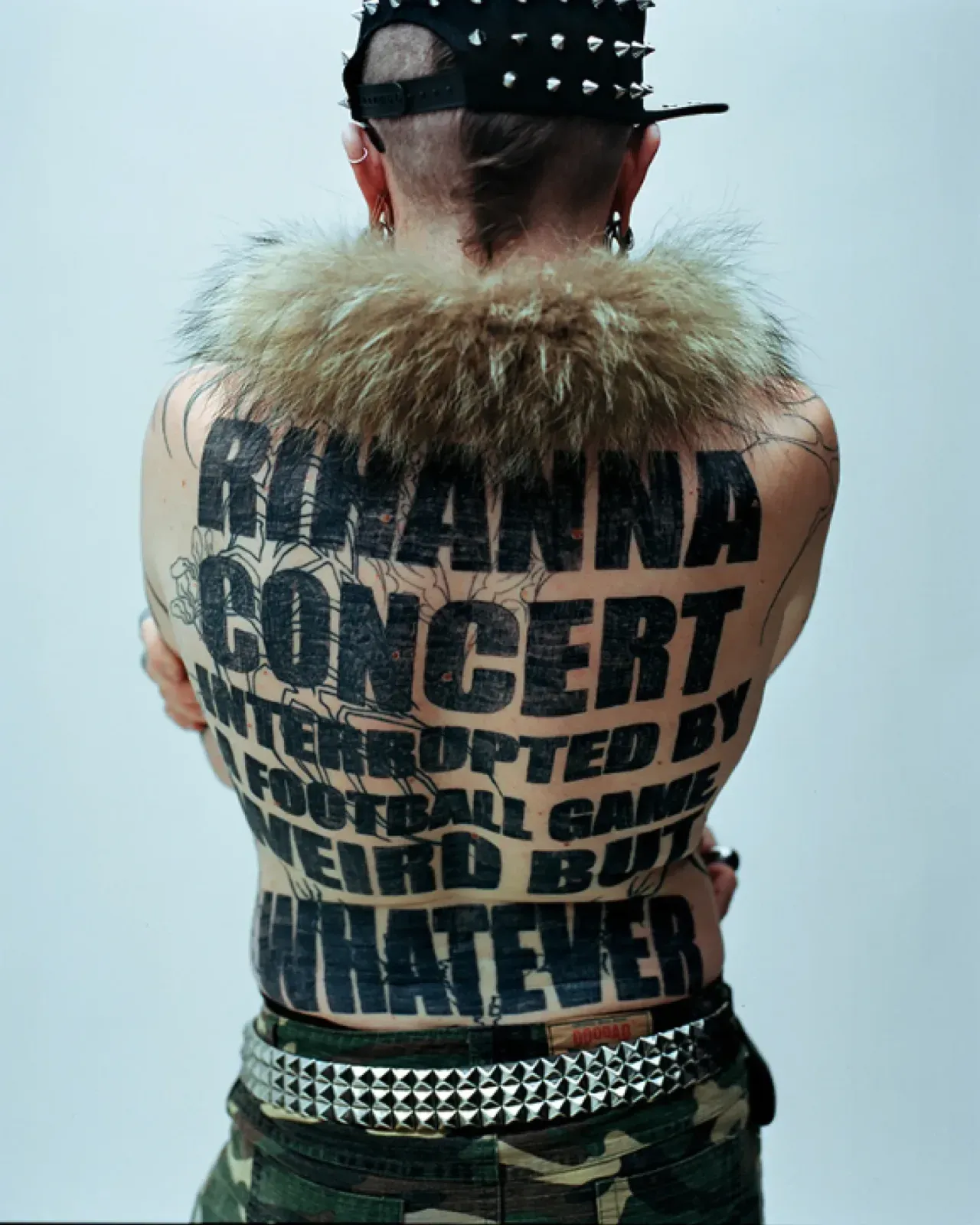 털 목도리를 두른 뒷모습. 등 전체에 「RIHANNA CONCERT INTERRUPTED BY A FOOTBALL GAME WEIRD BUT WHATEVER」 문신, 허리에 스터드 벨트