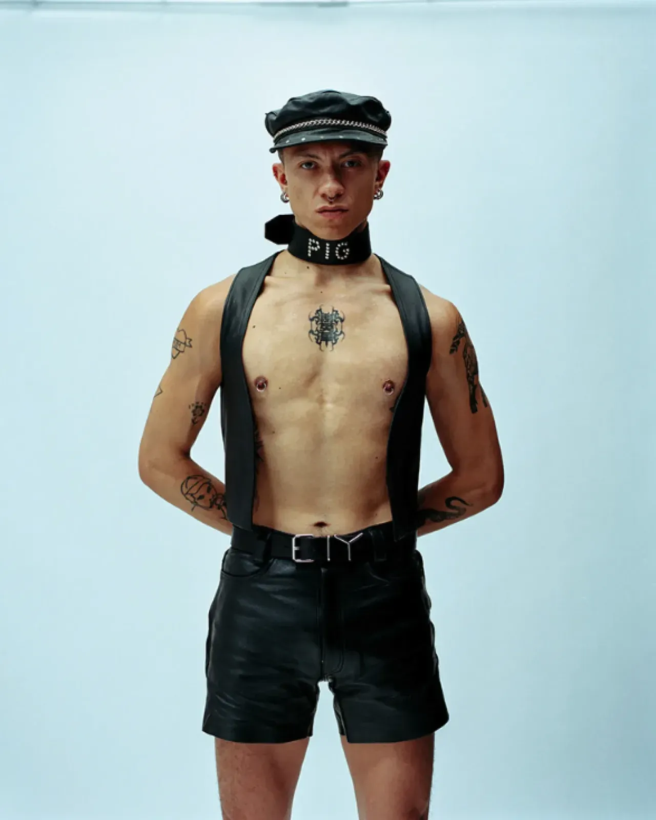 가죽 캡에 「PIG」라고 박힌 목줄, 맨가슴에 가죽 멜빵과 반바지를 입은 사람. 가슴 가운데에 문신