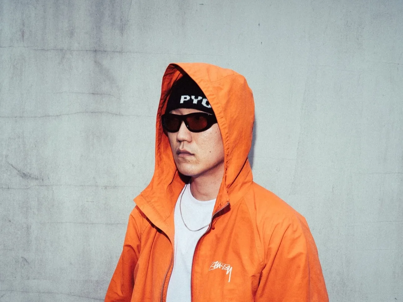 주황색 후드 집업과 선글라스를 쓰고 콘크리트 벽 앞에 선 DJ POOL