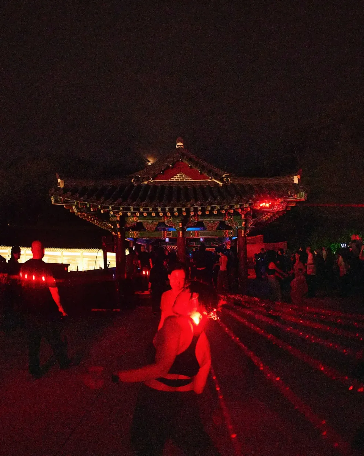 밤의 한옥 정자가 붉게 물들어 있고, 앞쪽 흙바닥에 붉은 레이저 줄기가 길게 깔린다. 가운데 사람이 몸을 낮추고 있다