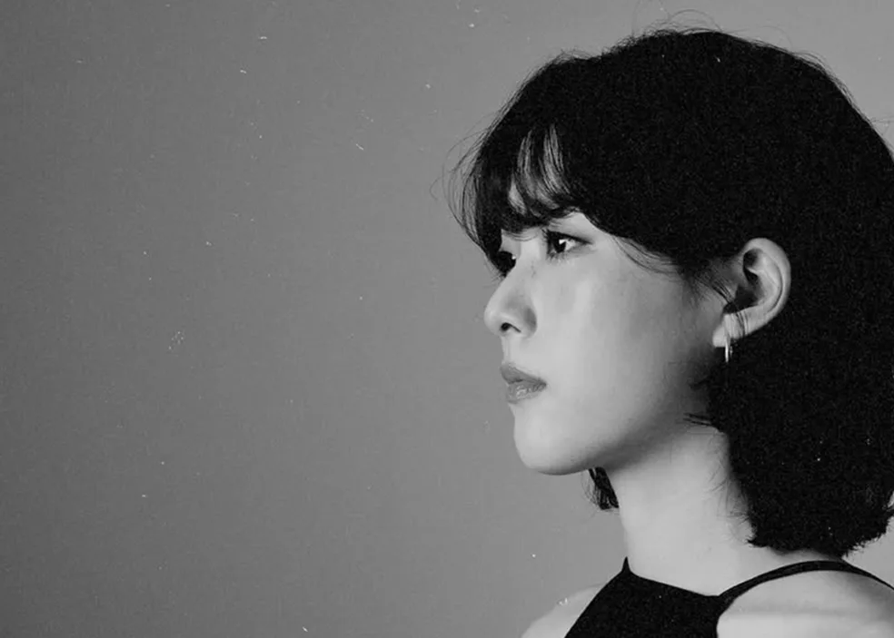 E3. 흑백 인물 사진. 귀걸이를 한 단발머리의 E3가 회색 배경 앞에서 옆을 보고 있다
