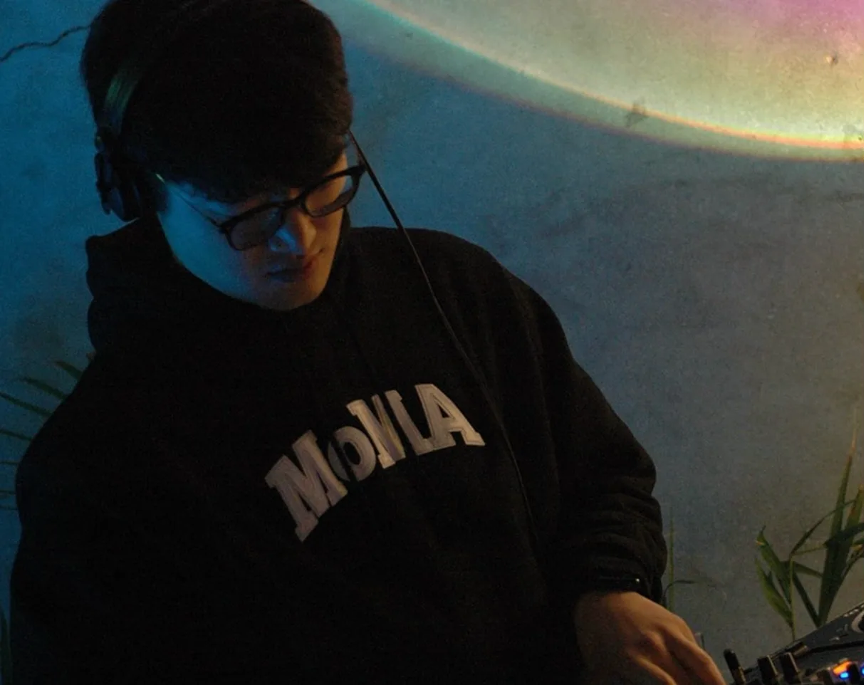 헤드폰을 걸고 안경을 쓴 판베씨가 고개를 숙인 채 DJ 믹서를 만지는 모습. MoMA 후드, 뒤로 무지개빛 조명
