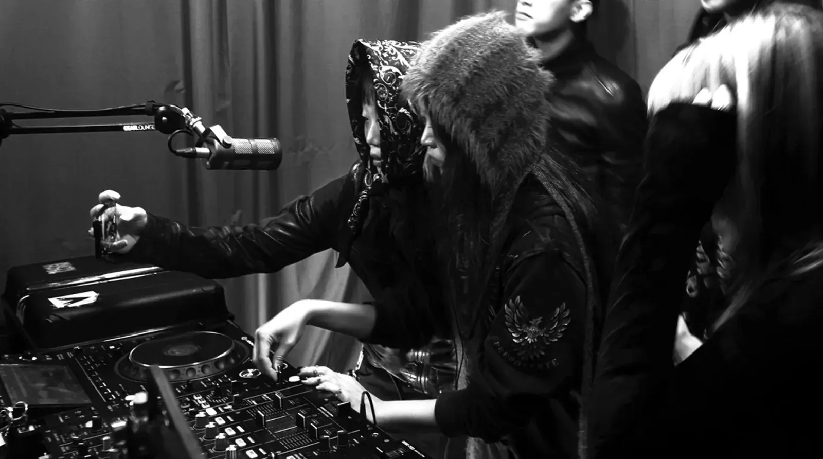 스카프와 털모자를 쓴 두 사람이 나란히 서서 DJ 믹서를 조작하고, 뒤에서 사람들이 지켜보는 흑백 사진