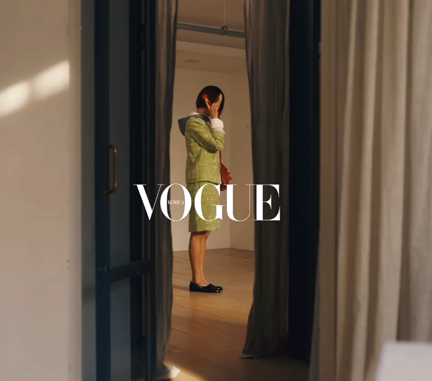 연두색 트위드 투피스를 입고 문틈 너머 방에서 통화하는 인물. 화면 가운데 VOGUE KOREA 로고