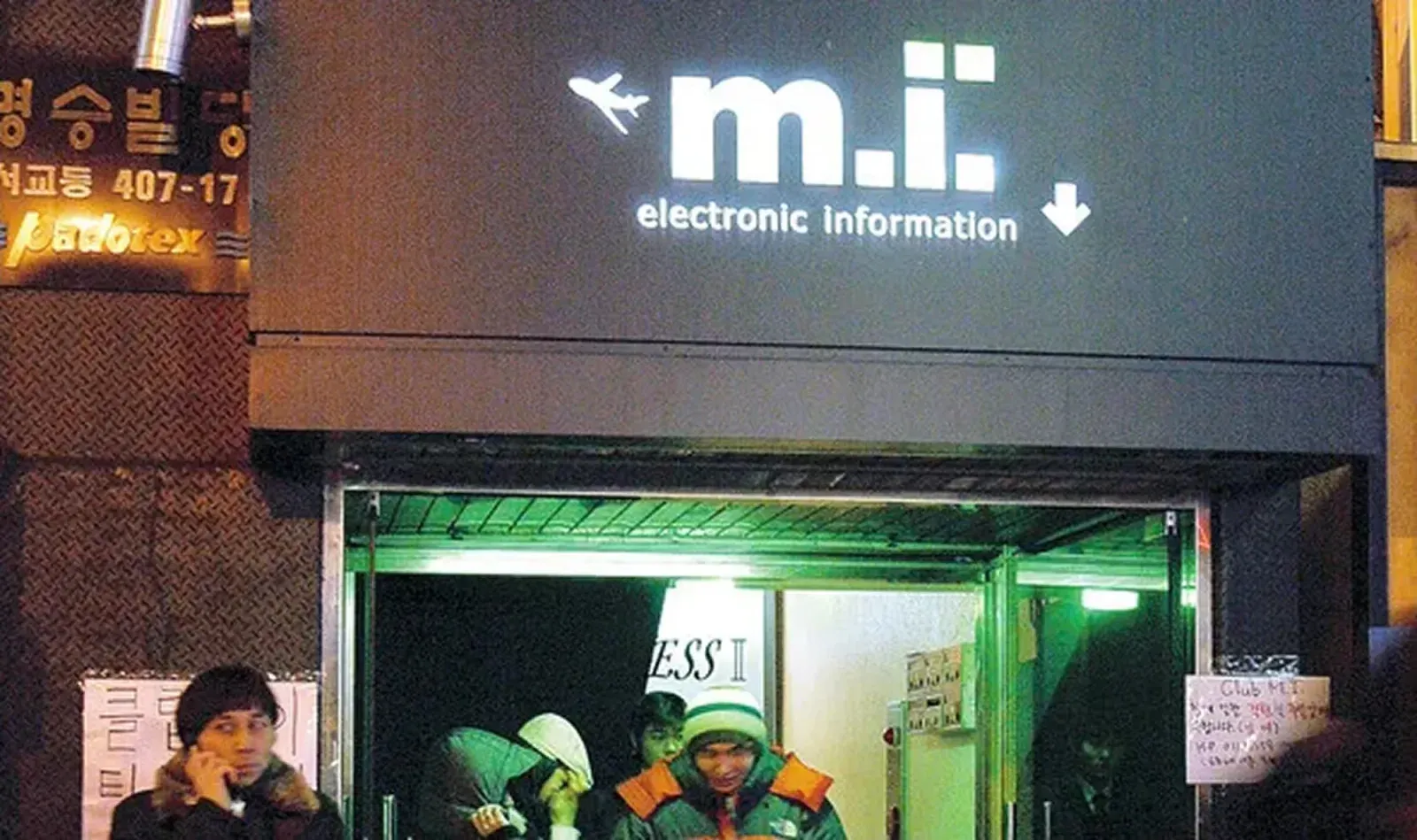 밤의 클럽 입구. 검은 간판에 흰 글자 「m.i. electronic information」과 비행기·화살표 그림, 열린 셔터 안에 사람들, 옆 벽에 「클럽 M.I」 안내문
